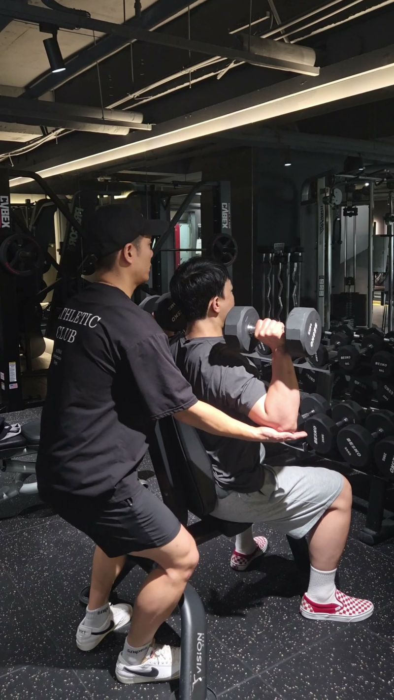 Trainer Kim Sanghyun holding a member's elbow in place during a dumbbell shoulder press